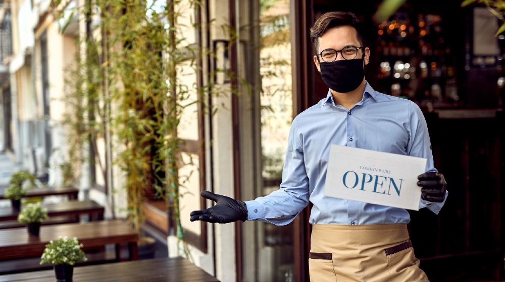 Happy waiter with protective face mask holding open sign while standing at cafe doorway-new ppp loans for small business-ss-featured