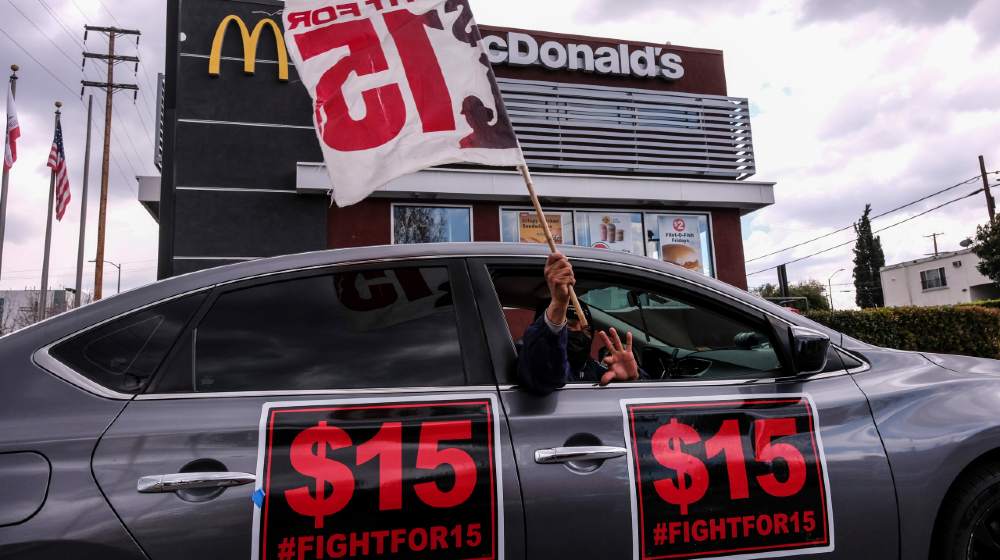 A worker holding a flag in a car during a rally to demand that four McDonald's workers who were fired after raising COVID-19 concerns get back pay and reinstatement | McDonald’s To Raise Wages For Corporate Store Workers | Featured