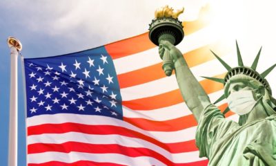 Statue of Liberty wearing a surgical mask, US american flag background | US COVID-19 Cases At Lowest Levels Since June 2020 | Featured