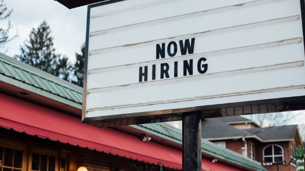 Typical american now hiring sign in black letters over the white background with local small business building | Montana To End Extra Jobless Benefits Due to Worker Shortage | featured