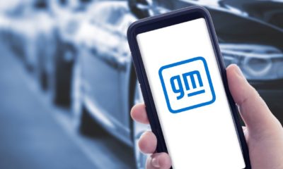 Hand holding a smart phone with new General Motors logo on screen and blurry cars on background | GM To Boost Spending On Electric and Self-Driving Cars | featured