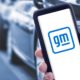 Hand holding a smart phone with new General Motors logo on screen and blurry cars on background | GM To Boost Spending On Electric and Self-Driving Cars | featured