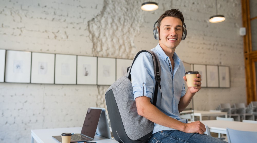 young handsome man sitting on table in headphones with backpack in co-working office | Walmart Offers Workers Free College Tuition And Books | featured