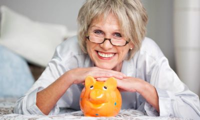 Conceptual portrait of happy senior female lying on bed with piggybank | The Best Retirement Income For 2021 | featured