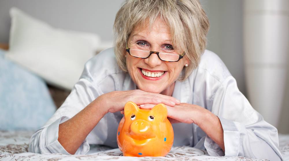 Conceptual portrait of happy senior female lying on bed with piggybank | The Best Retirement Income For 2021 | featured