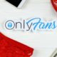Onlyfans paper logo with dollar bills, red mask and red lingerie on white wooden table | OnlyFans Reverses Course, Won’t Ban Explicit Content | featured