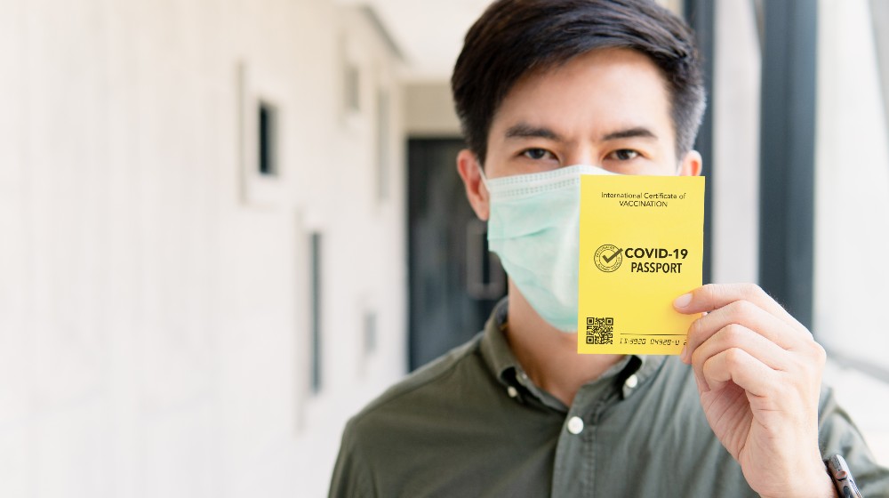 Portrait of a handsome asian man showing a risk-free certification COVID-19 passport to proof that he has been vaccinated against coronavirus and safe to travel | Are Vaccine Mandates Legal For Travelers? | featured