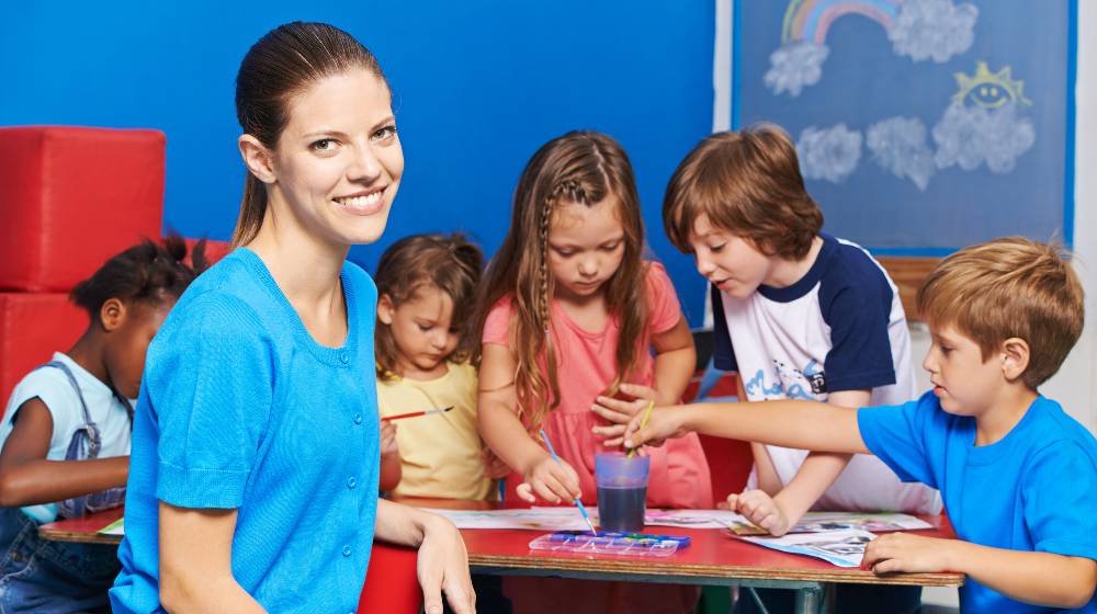 Woman as nursery teacher in kindergarten with group of children painting at table | Child Care Workers Are Leaving Their Jobs For Better Ones | featured