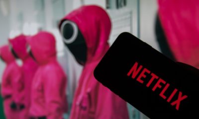 The Netflix logo on the display of a smartphone in front of a television with the new series Squid Game | Netflix’s Squid Game Cost $24m to Make, Now It’s Worth $900m | featured