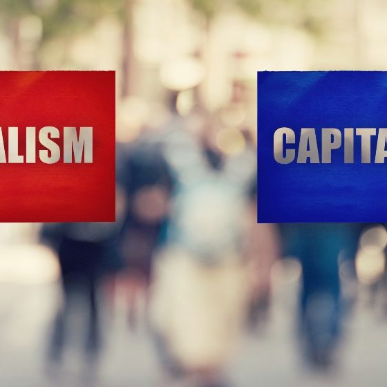 Two hands holding different colored paper sheet as socialist centralized economic planning versus capitalist liberated free market over crowded street | The Capitalism Vs Socialism Debate Remains Alive and Well | featured