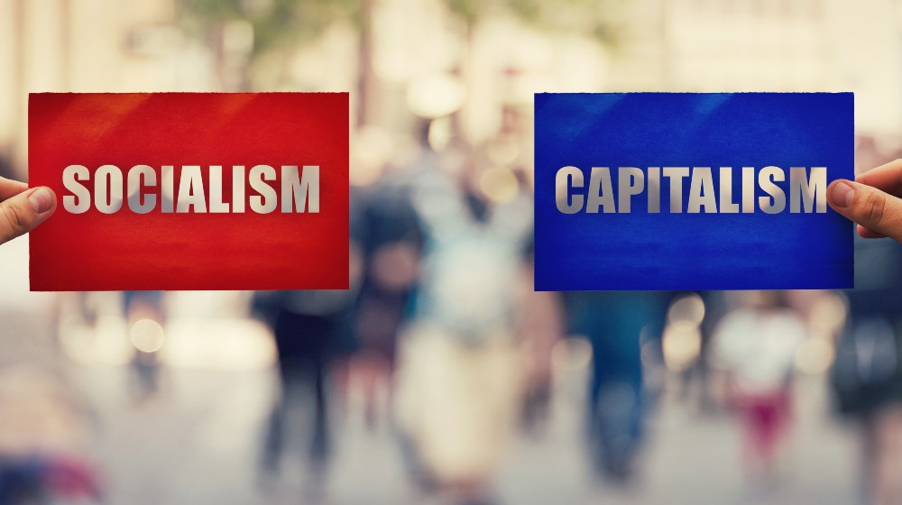 Two hands holding different colored paper sheet as socialist centralized economic planning versus capitalist liberated free market over crowded street | The Capitalism Vs Socialism Debate Remains Alive and Well | featured