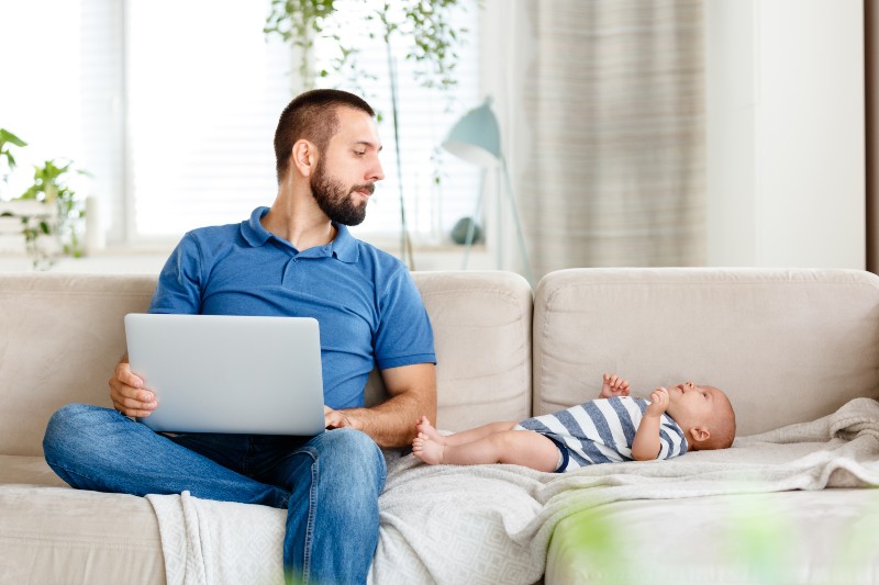 Young man sitting on sofa at home and using laptop while his baby boy lying next to him-Paternity Leave