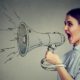 Young woman screaming in megaphone isolated on gray wall background | Facebook Whistleblower Reveals Identity In Interview | featured