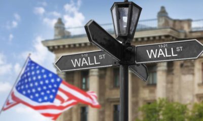 Wall Street signboard and stock exchange building on background | US Stock Market Posts New Record Highs On Strong Retail Earnings | featured