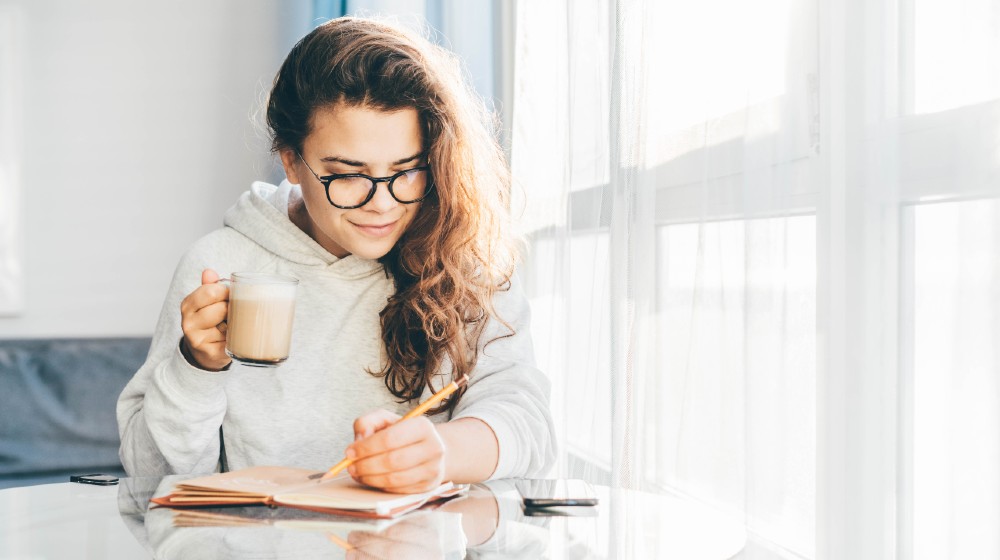 Woman writing notes in paper notebook.Woman sitting at the table with cup of coffee | 2 Hours in the Morning Will Put You in 2% Club! | featured