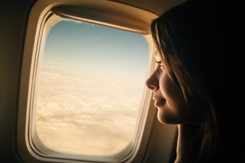Young woman looking through window in airplane | Airline CEOs Urge Biden To Drop Mask Requirements