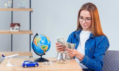 Young beautiful girl counts money from a piggy bank while sitting at a table | More States Want Students To Take Personal Finance Classes | featured