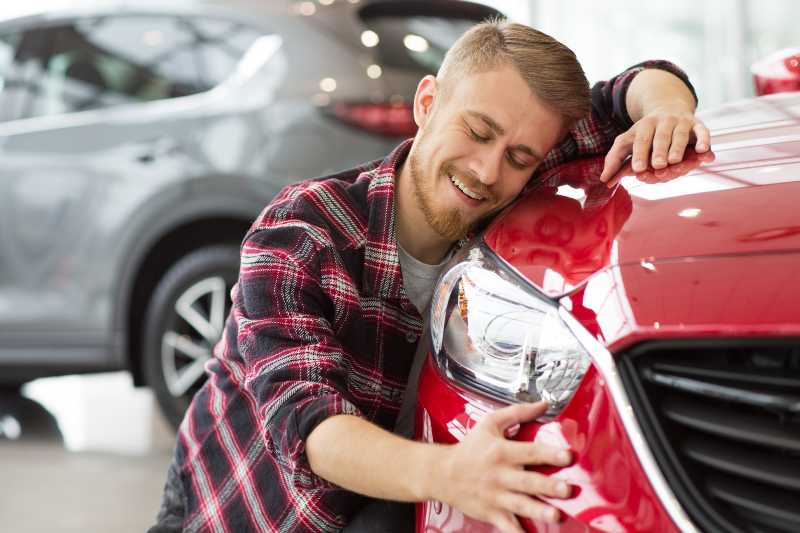 Shot of a handsome young happy bearded man embracing his new car at the dealership | Should We Buy a New Car?