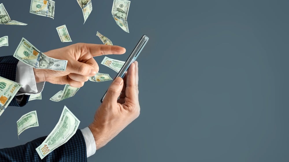 A hand from a smartphone transfers money and dollars fly out | US Still Undecided On Whether To Create Digital Currency | featured