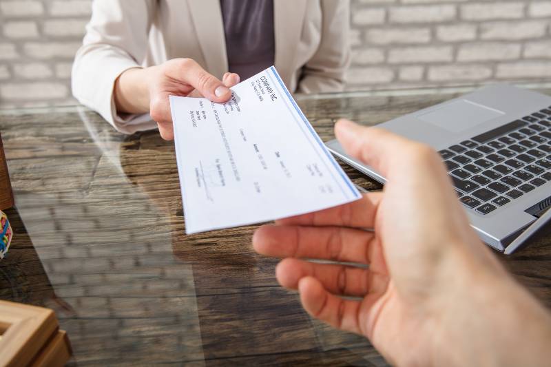 Close-up Of A Business Woman Giving Cheque To Her Colleague At Workplace In Office | Inflation Is Pushing Americans To Live Paycheck to Paycheck 