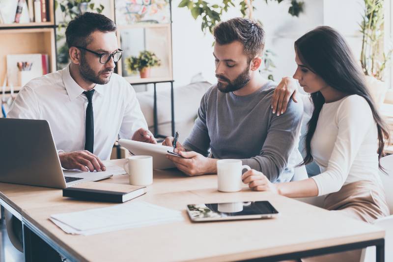 Confident young man signing some document while sitting together with his wife and man in shirt and tie-sell your company