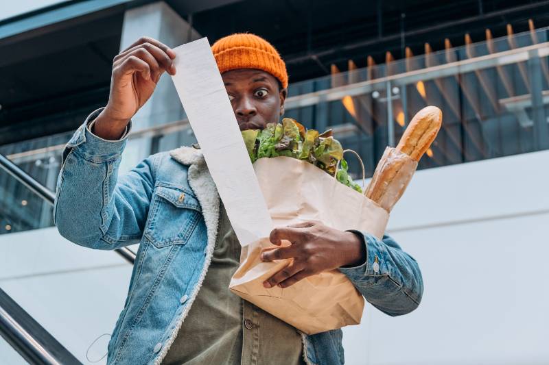 Doubting African-American person in denim jacket looks at sales paper receipt | American Households