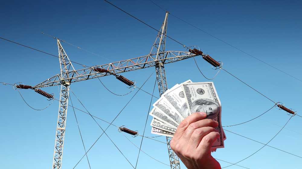 Hand holding US dollar bills in front of a high voltage power lines | Biden Signs $1 Trillion Infrastructure Bill Into Law | featured