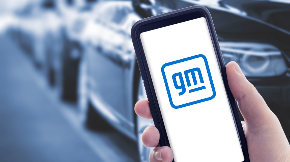 Hand holding a smart phone with new General Motors logo on screen and blurry cars on background | GM To Boost Spending On Electric and Self-Driving Cars | featured