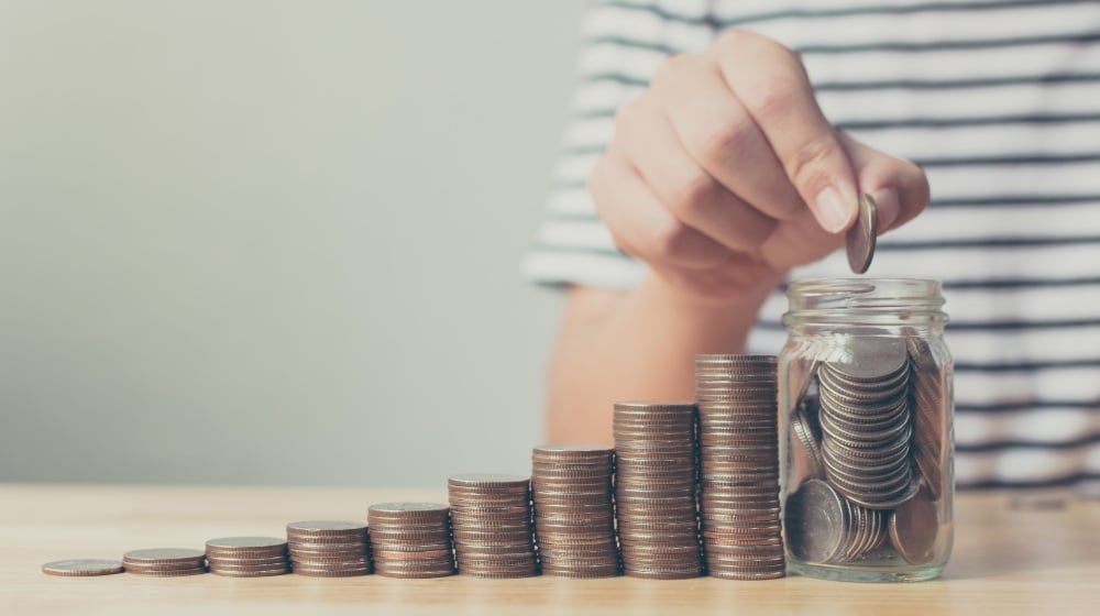 Hand of male putting coin in jar with money stack step-Money Goals-SS-Featured
