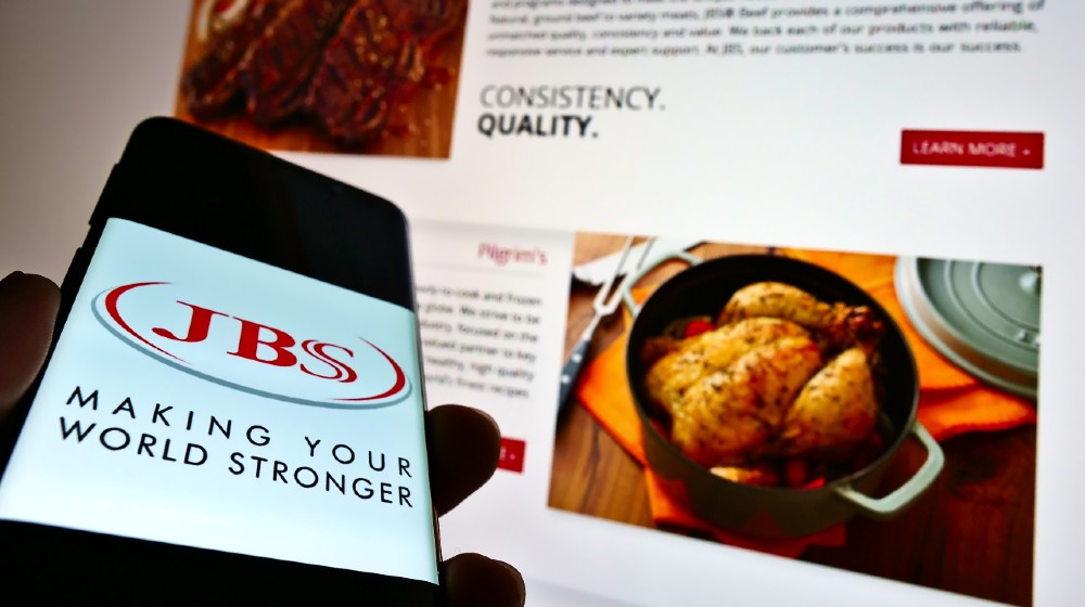 Person holding cellphone with logo of Brazilian meet processing company JBS S.A. on display with website | Cyberattack Hits World’s Largest Meat Supplier | Featured