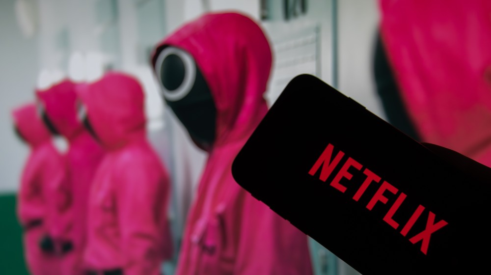 The Netflix logo on the display of a smartphone in front of a television with the new series Squid Game | Netflix’s Squid Game Cost $24m to Make, Now It’s Worth $900m | featured