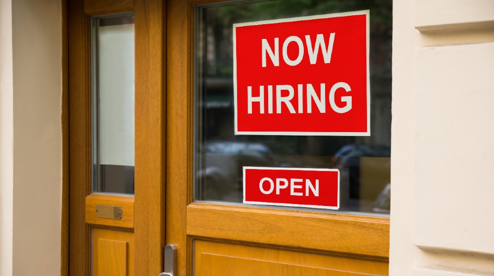 The Text Now Hiring Sticker Attached On Glass Door Of The Office | April Jobs Report Goes Bust Due to Worker Shortage | Featured