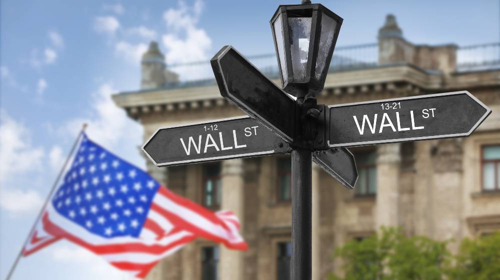 Wall Street signboard and stock exchange building on background | US Stock Market Posts New Record Highs On Strong Retail Earnings | featured