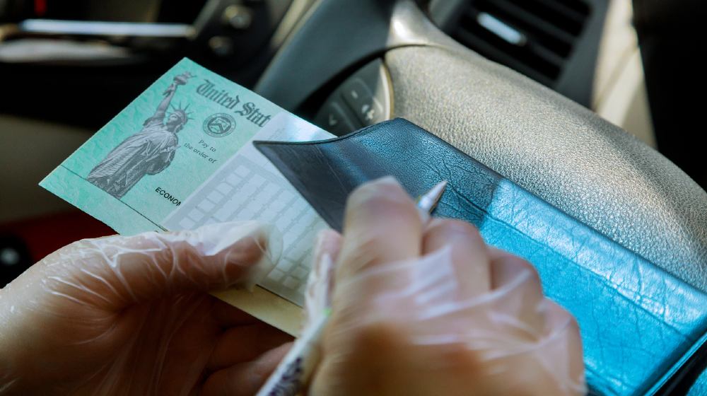Writing on blank check to with in car with gloves on depositing government stimulus check with gloves on for safety-Stimulus Money-ss-featured