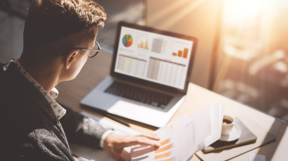 Young-finance-market-analyst-in-eyeglasses-working-at-sunny-office-on-laptop-while-sitting-at-wooden-table | How to Buy Private Stock | featured