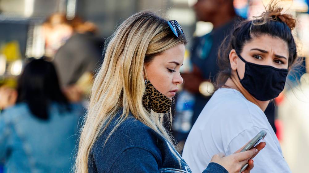 Young long hair blonde woman texting and walking with face mask down and mother friend looking back frustrated during covid-19 coronavirus pandemic | More Workers Support Heavier Penalties for Unvaccinated Ones | featured