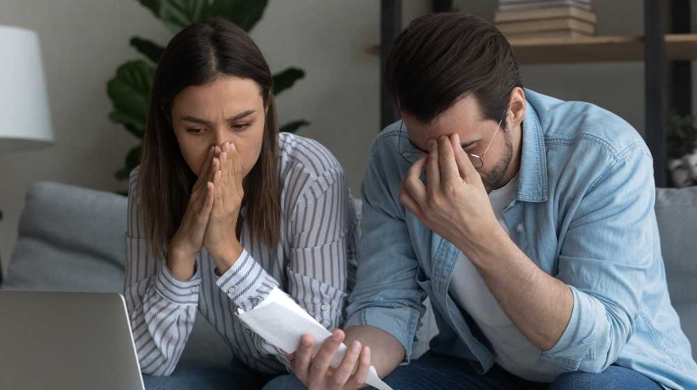 Young married couple sit on couch at home calculating expenses feel upset | US Household Expenses Are Now Higher By $296 Per Month | featured