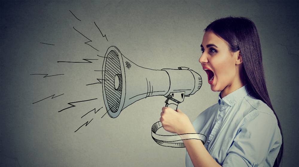 Young woman screaming in megaphone isolated on gray wall background | Facebook Whistleblower Reveals Identity In Interview | featured