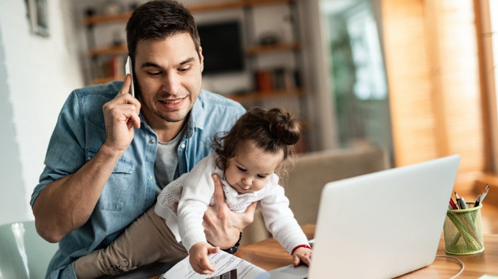 Young working father talking on the phone while babysitting his playful daughter at home-Work From Home-ss-featured