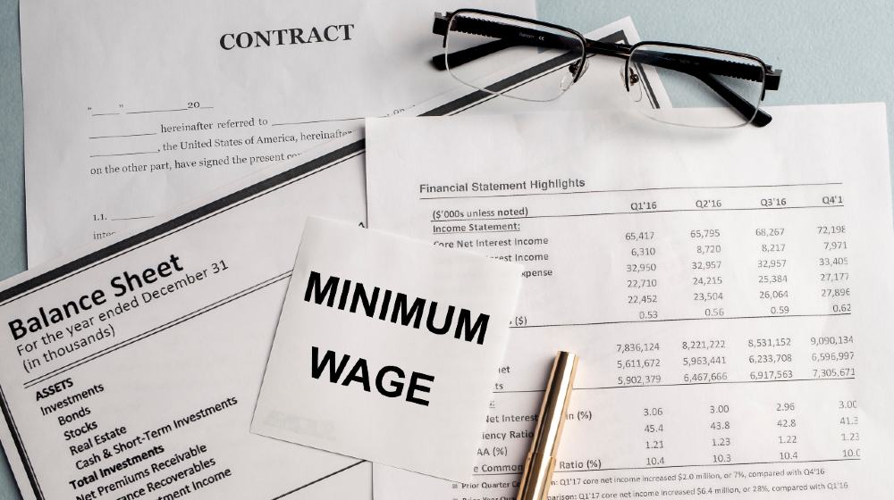a paper with MINIMUM WAGE text on backgrounwith documents and glasses | Biden To Sign $15 Minimum Wage For Government Contractors | Featured