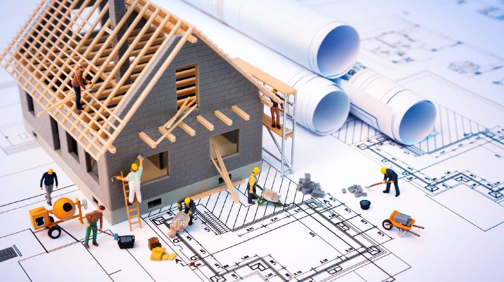 building house on blueprints with worker - construction project | House Construction Costs Are Going Through The Roof | Featured
